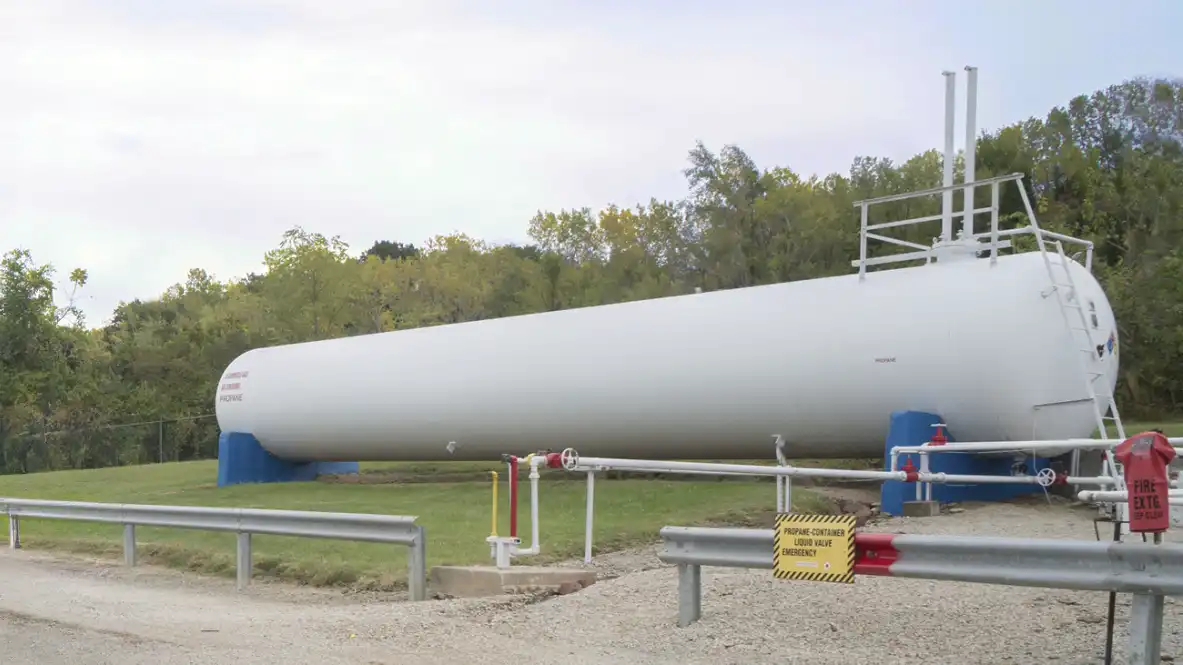 A propane tank in a grassy area, behind a guardrail. There are trees behind the grassy area.