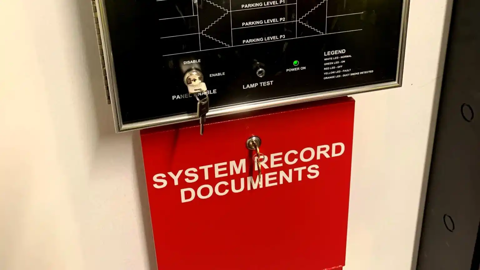 A smoke control system panel and the system record documents box that is on the wall underneath it. The document box is bright red with white lettering.