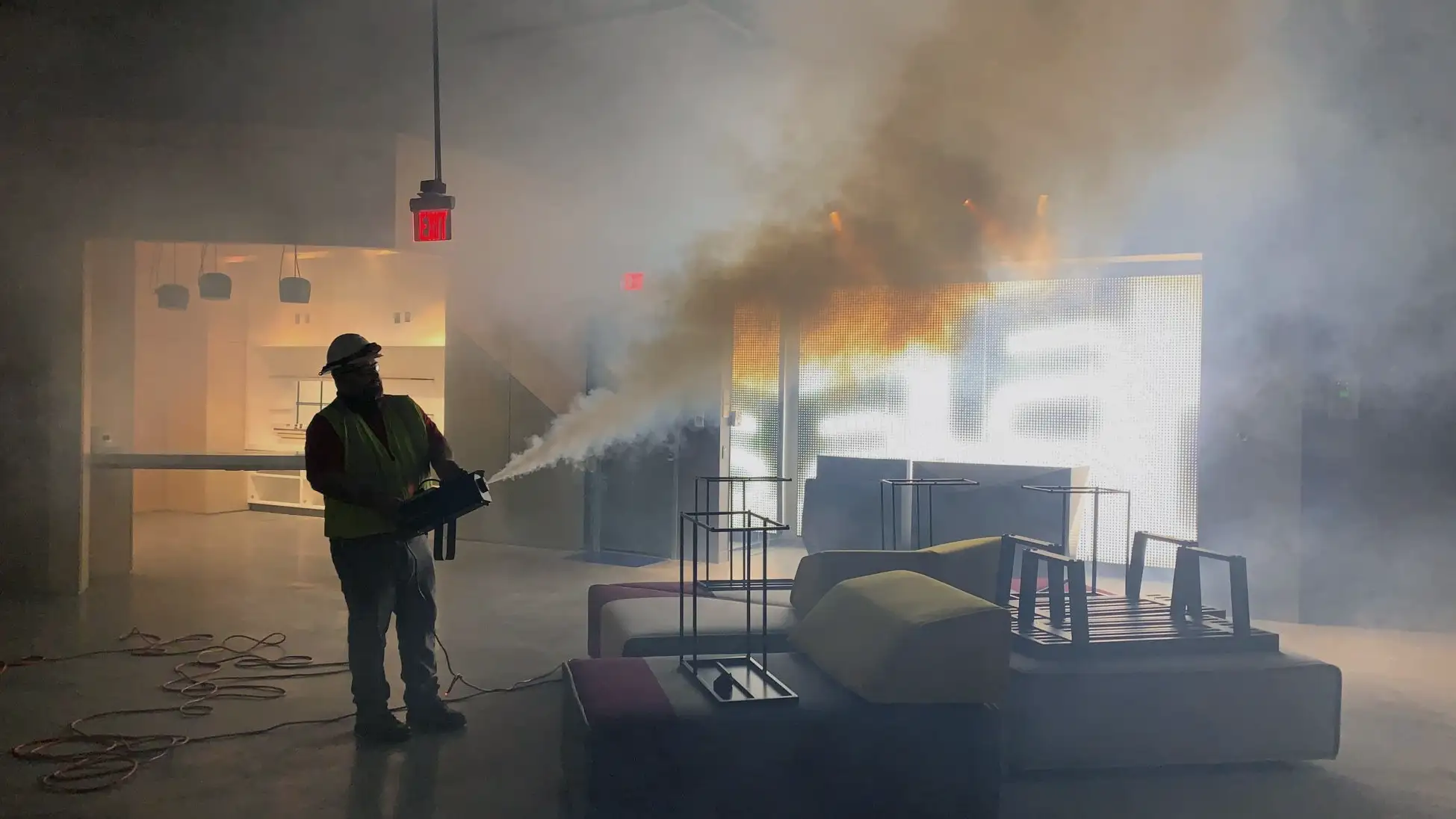 A worker in a yellow vest filling a room with smoke from a smoke machine in order to test the smoke control system. The room is dark and hazy from the smoke.